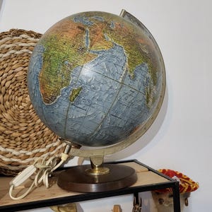 Könnte beinhalten: Ein beleuchteter Vintage-Globus mit einer detaillierten Weltkarte, blauen Ozeanen und bunten Landmassen. Der Globus ist auf einem runden Sockel aus dunklem Holz mit einem metallischen Meridianring montiert und besitzt ein weißes Stromkabel.