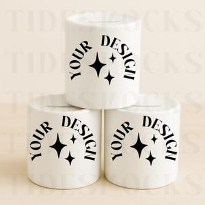 Puede incluir: Tres huchas de cerámica blanca con una ranura en la parte superior. Cada hucha tiene las palabras "YOUR DESIGN" en semicírculo sobre un diseño de estrella negra. Las huchas están apiladas unas sobre otras.