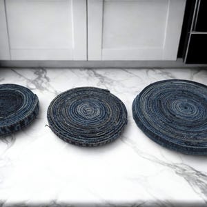 May include: Three circular denim coasters of varying sizes are arranged on a marble countertop. The coasters are made from rolled denim fabric, showcasing a spiral pattern. The denim is a mix of blue hues.