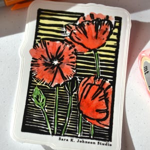 May include: A rectangular sticker with a floral design featuring red poppies with black and yellow accents. The sticker has a white border and the text "Sara K. Johnson Studio". An orange Sharpie marker is visible in the background.