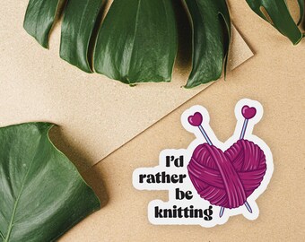 May include: A white sticker with a pink heart-shaped ball of yarn and two knitting needles. The text "I'd rather be knitting" is written in black below the yarn.