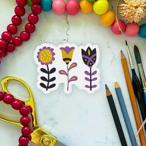 Puede incluir: Tres pegatinas de flores coloridas con detalles en morado, amarillo y blanco. Las pegatinas están sobre un fondo blanco con un par de tijeras doradas y una cadena de cuentas rojas.