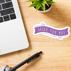 May include: A purple sticker with the text "Seize the Day" and "Epilepsy Awareness" in a banner design. The sticker is on a wooden desk next to a laptop, a pen, and a small potted plant.