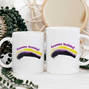 Può includere: Due tazze in ceramica bianca con un design viola, nero e giallo e il testo "Assume Nothing".