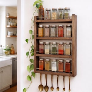 May include: A wooden spice rack with multiple shelves and small glass jars filled with various spices. The rack has brass accents and hanging brass measuring spoons. A trailing green plant adds a natural touch to the kitchen decor.