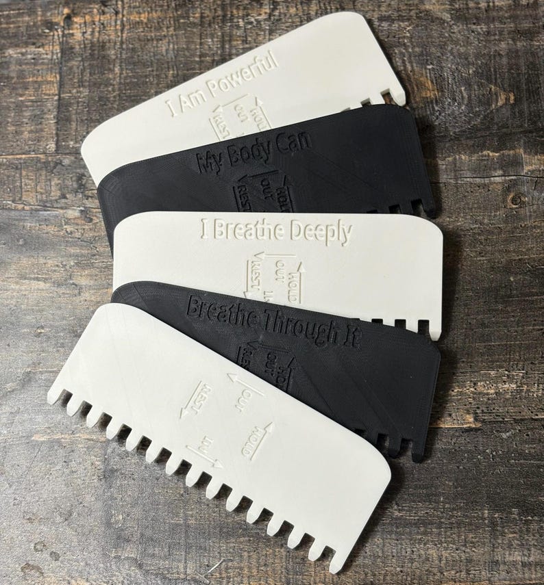 May include: A stack of white and black plastic tools with a comb-like edge. Each tool has an inspirational phrase embossed on it, such as "I Am Powerful" and "Breathe Through It." Arrows indicate the breathing process.
