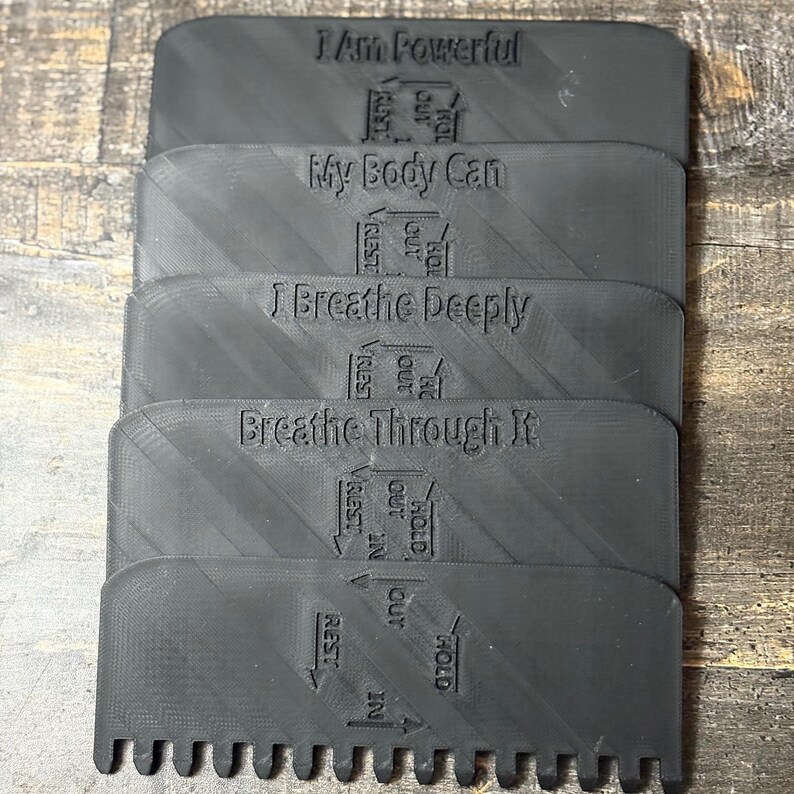 May include: Stacked black plastic meditation tools with text: "I Am Powerful", "My Body Can", "I Breathe Deeply", and "Breathe Through It". Each tool has a comb-like edge and directional arrows for breathwork. The tools are designed for mindfulness practices.
