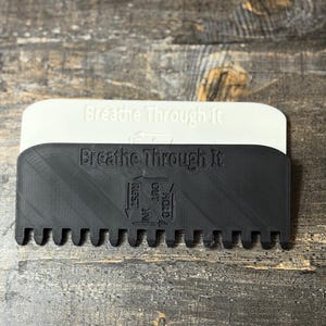 May include: A black and white 3D-printed breathing tool. The black tool has a comb-like edge and the text "Breathe Through It" and directional arrows. The white tool on top also has the text "Breathe Through It".
