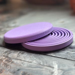 May include: A lavender-colored, circular object with a flat top and a ridged, spiral design on the inside. The text "Simply Loved Doula" is embossed on the side. The object is made of a solid material and sits on a textured surface.