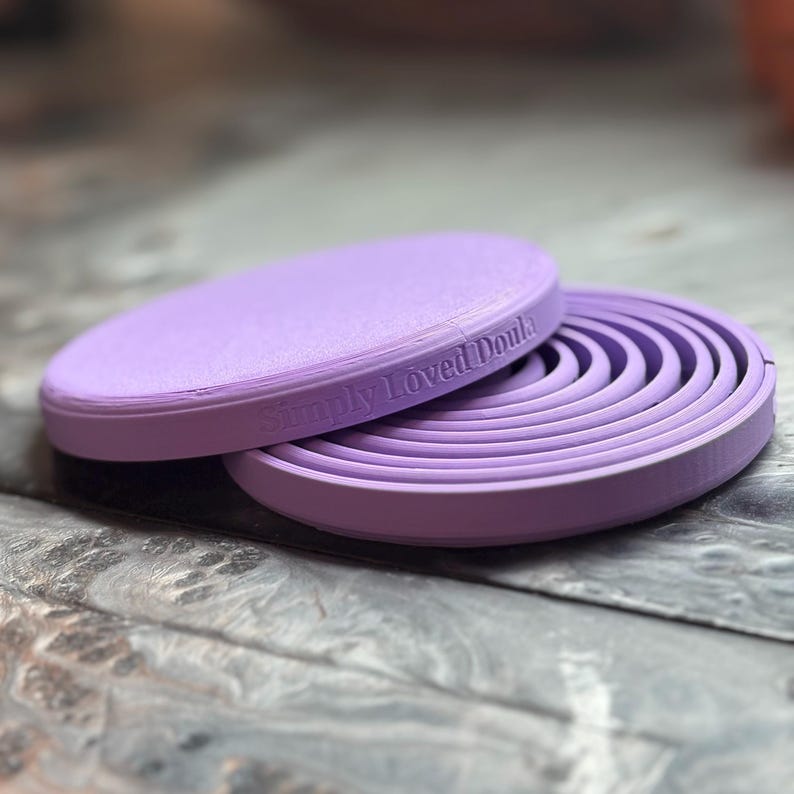 May include: A lavender-colored, circular object with a flat top and a ridged, spiral design on the inside. The text "Simply Loved Doula" is embossed on the side. The object is made of a solid material and sits on a textured surface.
