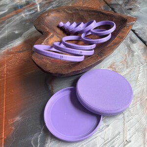 May include: A collection of light purple ring-shaped tools, numbered from 4 to 10, are arranged in a wooden bowl. Two circular containers, also light purple, are positioned below the bowl. The tools appear to be for crafting or measuring.