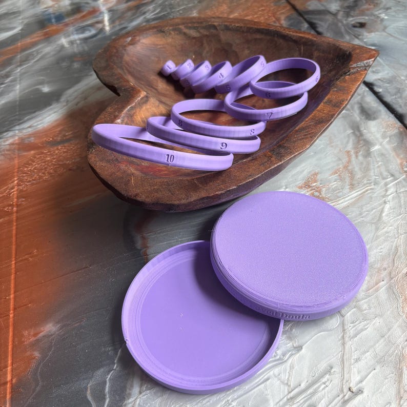 May include: A collection of light purple ring-shaped tools, numbered from 4 to 10, are arranged in a wooden bowl. Two circular containers, also light purple, are positioned below the bowl. The tools appear to be for crafting or measuring.