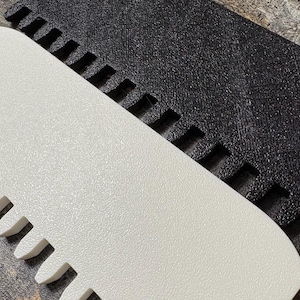 May include: Close-up of two textured, rectangular objects. One is white with a rounded edge and a series of evenly spaced, rectangular cutouts. The other is black with a similar texture and cutouts. The objects are angled towards each other.