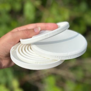 Puede incluir: Producto blanco, circular, impreso en 3D con el texto "My Body Can". El producto tiene múltiples capas, con el número "10" visible en una capa y el número "9" en otra. El producto se sostiene en una mano.