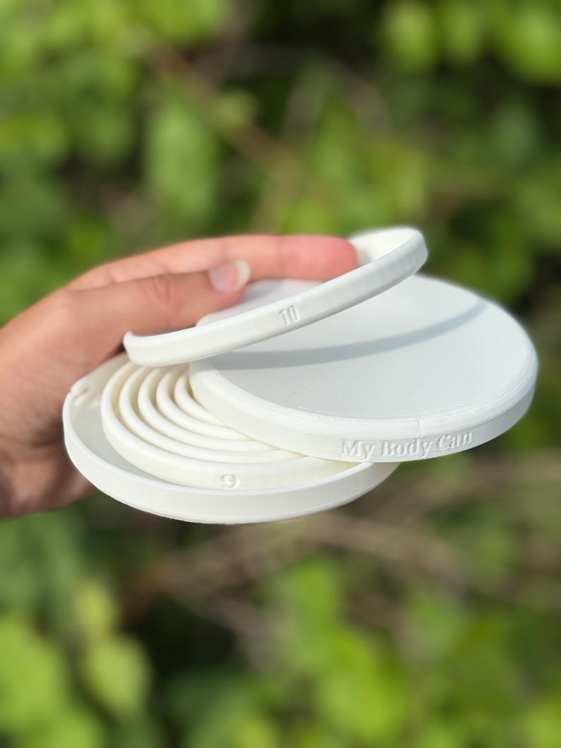 May include: White, circular, 3D-printed product with the text "My Body Can". The product has multiple layers, with the number "10" visible on one layer and the number "9" on another. The product is held in a hand.