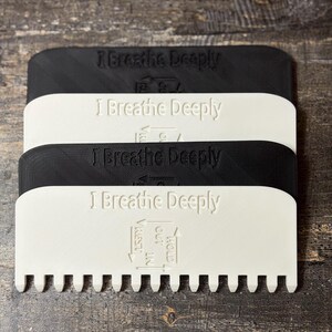 May include: Four rectangular, paddle-shaped tools in black and white, stacked on top of each other. Each tool has the text "I Breathe Deeply" embossed on the top. The bottom two tools have a row of teeth along the bottom edge. The bottom tool also has the words "Hold Out In Rest" with arrows.