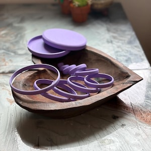 May include: A collection of lavender-colored baking tools, including round cookie cutters with numbers, and two lids, are arranged in a wooden bowl. The bowl sits on a textured, light-colored surface.