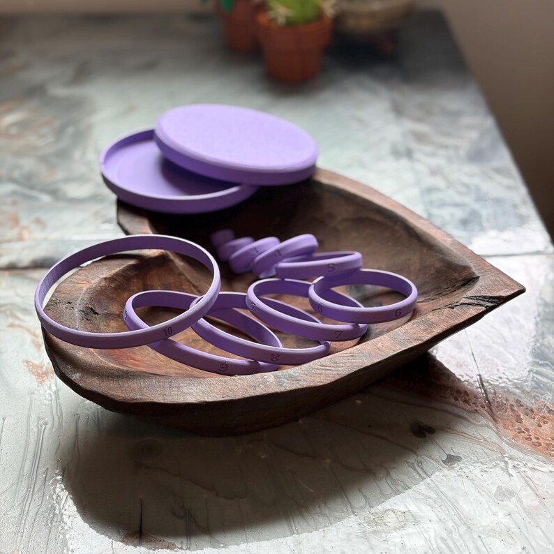 May include: A collection of lavender-colored baking tools, including round cookie cutters with numbers, and two lids, are arranged in a wooden bowl. The bowl sits on a textured, light-colored surface.