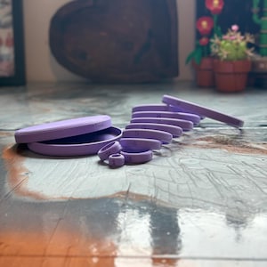 May include: A set of lavender-colored ring sizers, including a tray and individual rings numbered 1 through 9. The rings are arranged in a stack, with the tray open to the side. The text "Under Loved Doula" is visible on the tray.