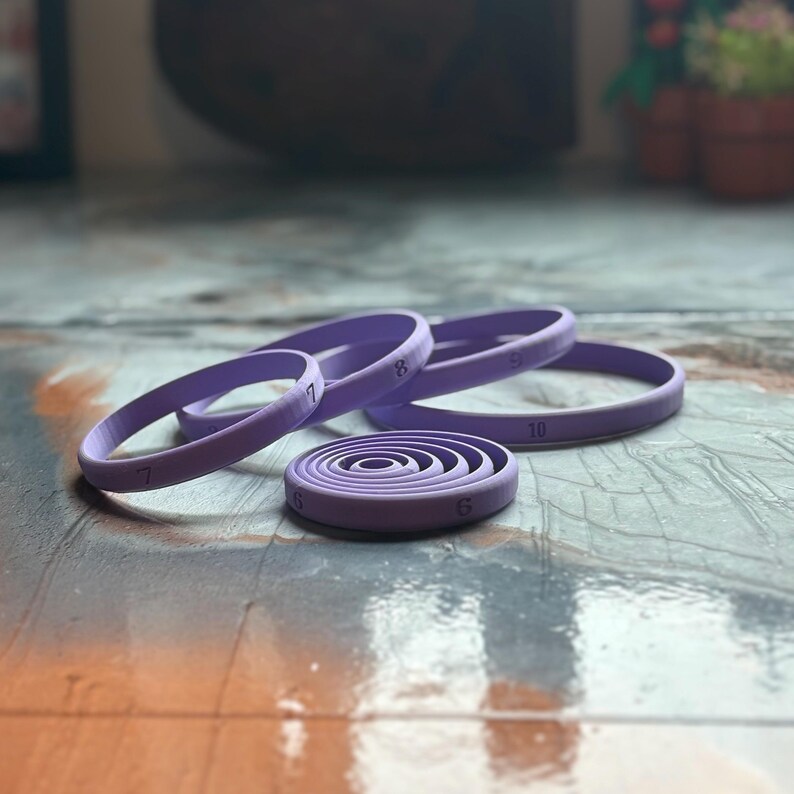 May include: A collection of purple, circular cookie cutters in various sizes. The cutters are numbered 6 through 10, with the smallest cutter in a spiral design. The cutters are made of a smooth material and are arranged on a textured surface.