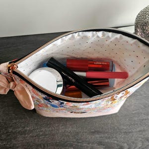 May include: A zippered pouch featuring a colorful character print on the exterior and a light-colored interior with small gold dots. The pouch is accented with a light pink fabric bow on the side and contains various cosmetic items like tubes and a jar.