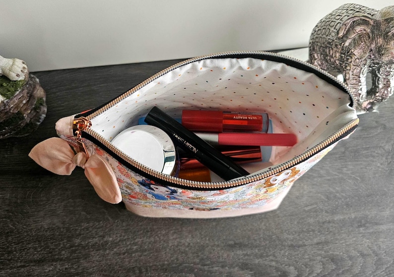 May include: A zippered pouch featuring a colorful character print on the exterior and a light-colored interior with small gold dots. The pouch is accented with a light pink fabric bow on the side and contains various cosmetic items like tubes and a jar.