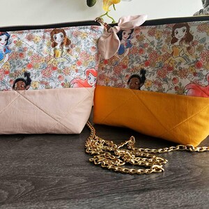 May include: Two quilted fabric pouches featuring a floral pattern with cartoon princess characters. One pouch has a light pink base and the other a mustard yellow base. The yellow pouch includes a gold-tone metal chain strap and a small fabric bow.