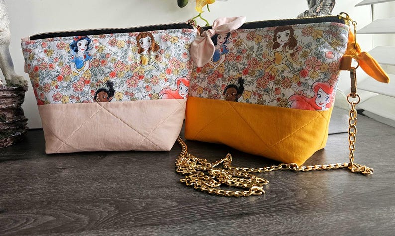 May include: Two quilted fabric pouches featuring a floral pattern with cartoon princess characters. One pouch has a light pink base and the other a mustard yellow base. The yellow pouch includes a gold-tone metal chain strap and a small fabric bow.