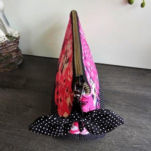 Peut inclure: Une pochette triangulaire zipp&eacute;e en tissu rose vif avec un motif floral et de personnages blancs. La pochette poss&egrave;de une base et des panneaux lat&eacute;raux en tissu &agrave; pois noirs et blancs, termin&eacute;s par une tirette en forme de n&oelig;ud &agrave; pois.