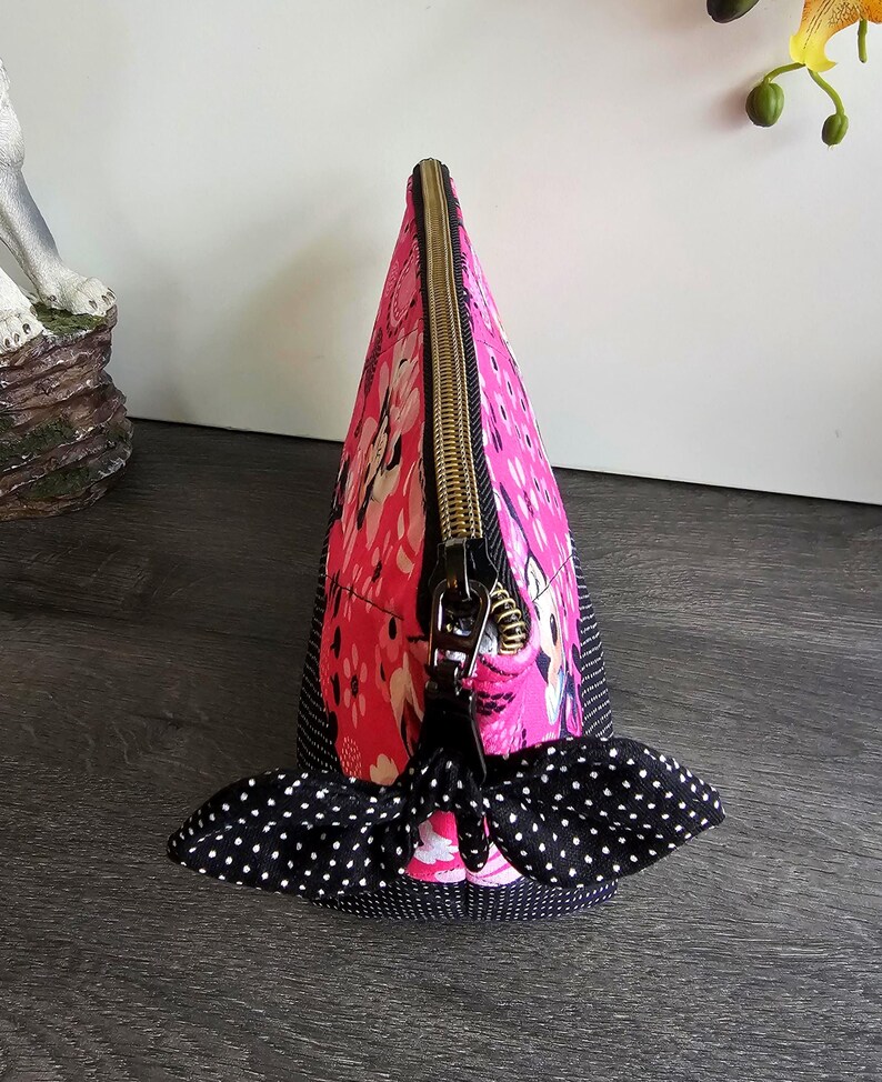 Peut inclure: Une pochette triangulaire zipp&eacute;e en tissu rose vif avec un motif floral et de personnages blancs. La pochette poss&egrave;de une base et des panneaux lat&eacute;raux en tissu &agrave; pois noirs et blancs, termin&eacute;s par une tirette en forme de n&oelig;ud &agrave; pois.
