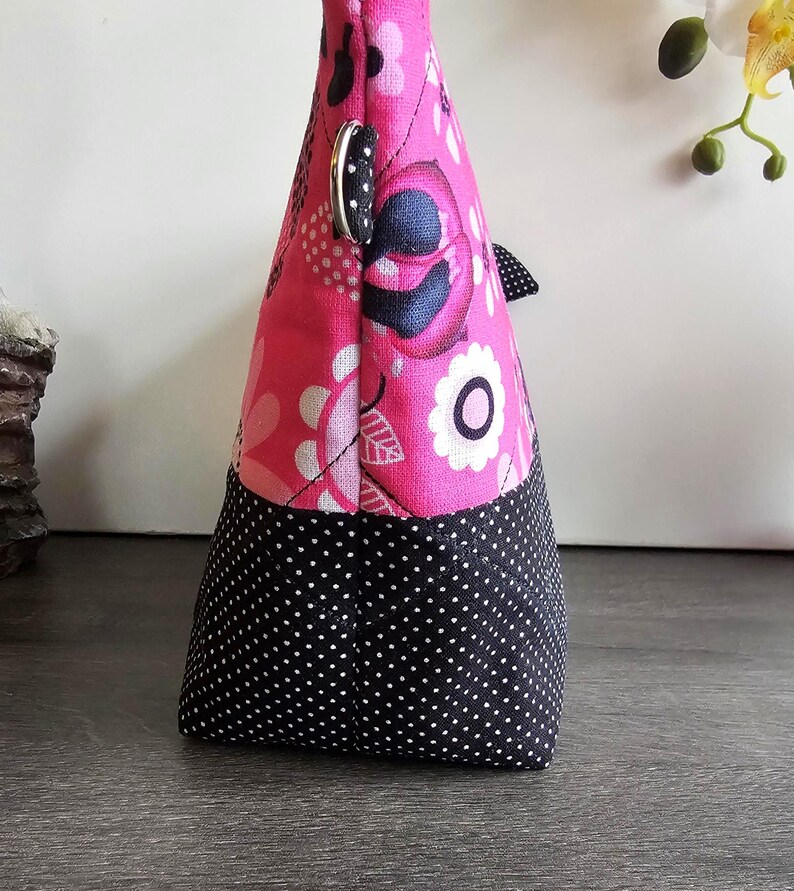 Peut inclure: Une pochette en tissu avec une section sup&eacute;rieure rose vif &agrave; motif floral noir et blanc et une section inf&eacute;rieure noire &agrave; pois blancs. Le sac comprend un anneau en D en m&eacute;tal argent&eacute; sur le c&ocirc;t&eacute; et des coutures matelass&eacute;es visibles.