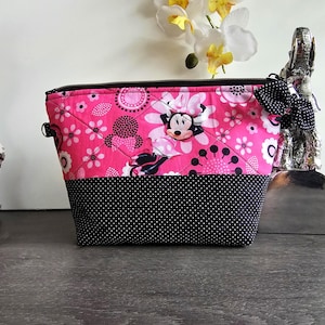Peut inclure: Une pochette zipp&eacute;e matelass&eacute;e pr&eacute;sentant un tissu rose vif avec un motif Minnie Mouse et floral sur la moiti&eacute; sup&eacute;rieure et un tissu noir &agrave; pois blancs sur la moiti&eacute; inf&eacute;rieure. Un n&oelig;ud &agrave; pois noir et blanc est attach&eacute; &agrave; la fermeture &eacute;clair.