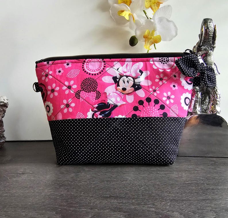 Peut inclure: Une pochette zipp&eacute;e matelass&eacute;e pr&eacute;sentant un tissu rose vif avec un motif Minnie Mouse et floral sur la moiti&eacute; sup&eacute;rieure et un tissu noir &agrave; pois blancs sur la moiti&eacute; inf&eacute;rieure. Un n&oelig;ud &agrave; pois noir et blanc est attach&eacute; &agrave; la fermeture &eacute;clair.