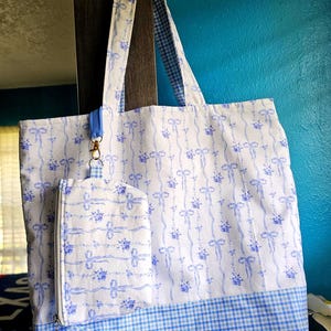 Puede incluir: Bolso tote blanco y juego de bolsa a juego con un patrón floral y de lazo azul. El bolso tote tiene ribetes y asas de cuadros azules. La bolsa está sujeta con un cierre dorado. La bolsa está colgada contra una pared azul.
