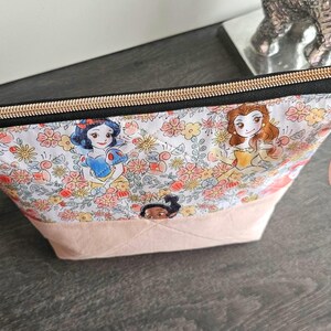 May include: A zippered pouch featuring a white fabric printed with colorful floral patterns and illustrations of various animated princesses. The bottom section is a solid light pink fabric, and the top closure has a black zipper with gold hardware.
