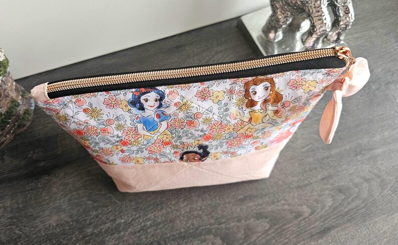 May include: A zippered pouch featuring a white fabric printed with colorful floral patterns and illustrations of various animated princesses. The bottom section is a solid light pink fabric, and the top closure has a black zipper with gold hardware.