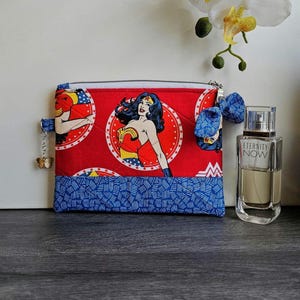 Puede incluir: Un estuche con cremallera rojo y azul con el tema de Wonder Woman con un dije de mariposa. El estuche presenta un gráfico de Wonder Woman y una parte inferior azul estampada. Un lazo azul y un frasco de perfume también están en la imagen.
