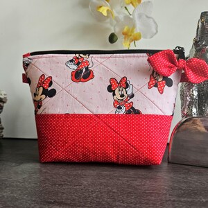 Pu&ograve; includere: Una pochette in tessuto trapuntato con sfondo rosa chiaro, un motivo ripetuto di Minnie Mouse e una sezione inferiore rossa a pois. La pochette presenta una chiusura a zip nera e un fiocco decorativo rosso a pois attaccato sul lato.