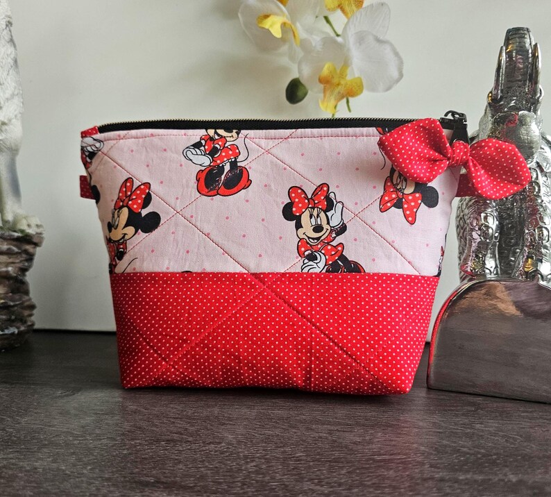 Pu&ograve; includere: Una pochette in tessuto trapuntato con sfondo rosa chiaro, un motivo ripetuto di Minnie Mouse e una sezione inferiore rossa a pois. La pochette presenta una chiusura a zip nera e un fiocco decorativo rosso a pois attaccato sul lato.