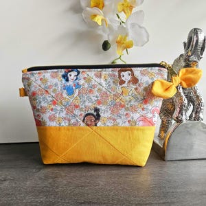 May include: A quilted zippered pouch featuring a white fabric top with a colorful floral pattern and illustrations of various princesses, paired with a solid mustard yellow fabric bottom. A yellow fabric bow is attached to the gold zipper pull.