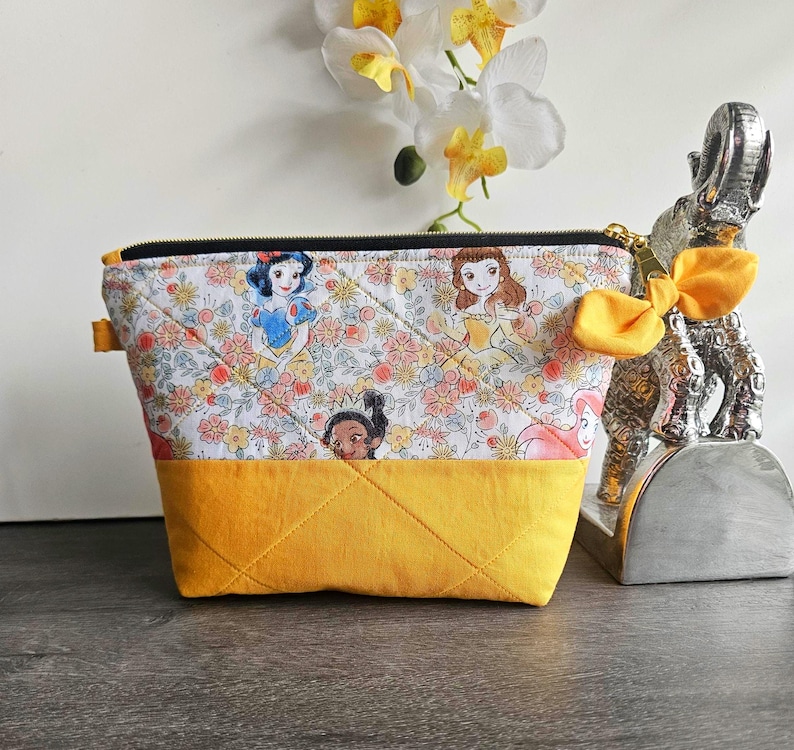 May include: A quilted zippered pouch featuring a white fabric top with a colorful floral pattern and illustrations of various princesses, paired with a solid mustard yellow fabric bottom. A yellow fabric bow is attached to the gold zipper pull.