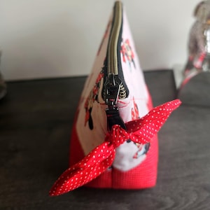 Pu&ograve; includere: Una pochette in tessuto triangolare con sfondo rosa chiaro e una stampa ripetuta di personaggi dei cartoni animati. La pochette presenta una base rossa tinta unita, una chiusura a zip in metallo nero e un tirante in tessuto rosso a pois.