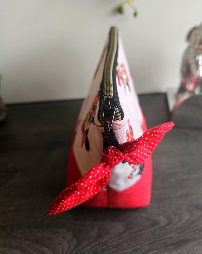 Pu&ograve; includere: Una pochette in tessuto triangolare con sfondo rosa chiaro e una stampa ripetuta di personaggi dei cartoni animati. La pochette presenta una base rossa tinta unita, una chiusura a zip in metallo nero e un tirante in tessuto rosso a pois.
