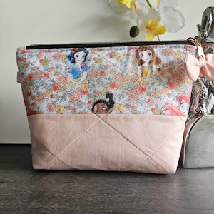 May include: A quilted zippered pouch featuring a top panel with a floral pattern and cartoon character illustrations, and a solid light pink bottom panel. A small light pink fabric bow is attached to the side near the black zipper closure.