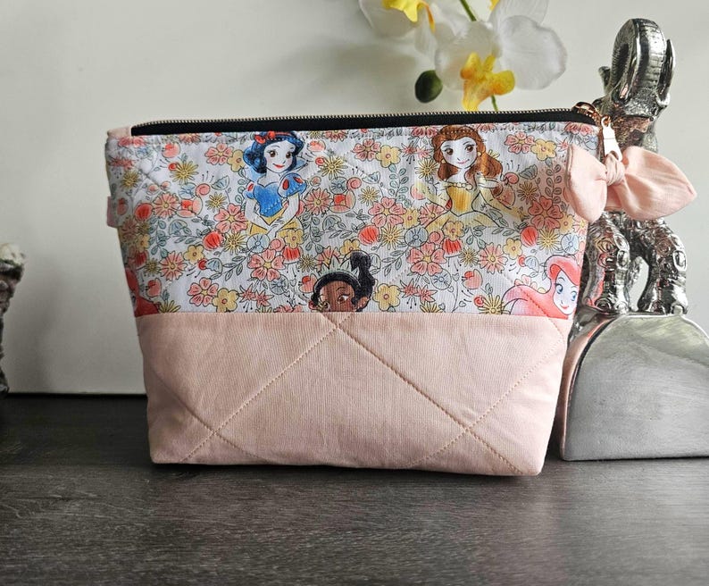 May include: A quilted zippered pouch featuring a top panel with a floral pattern and cartoon character illustrations, and a solid light pink bottom panel. A small light pink fabric bow is attached to the side near the black zipper closure.