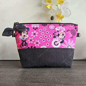 Peut inclure: Une pochette zipp&eacute;e matelass&eacute;e avec un fond rose vif orn&eacute; d'un motif r&eacute;p&eacute;titif de Minnie Mouse et de fleurs. La partie inf&eacute;rieure est noire &agrave; pois blancs. Un n&oelig;ud en tissu &agrave; pois noirs et blancs est fix&eacute; sur la fermeture &eacute;clair lat&eacute;rale.