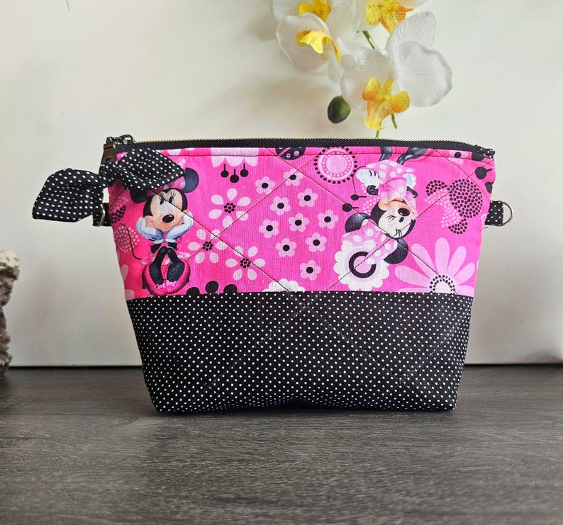Peut inclure: Une pochette zipp&eacute;e matelass&eacute;e avec un fond rose vif orn&eacute; d'un motif r&eacute;p&eacute;titif de Minnie Mouse et de fleurs. La partie inf&eacute;rieure est noire &agrave; pois blancs. Un n&oelig;ud en tissu &agrave; pois noirs et blancs est fix&eacute; sur la fermeture &eacute;clair lat&eacute;rale.