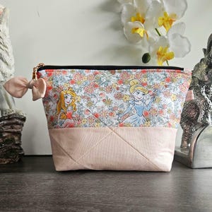 May include: A zippered pouch featuring a top section with a floral pattern and cartoon princess illustrations, and a solid light pink quilted bottom section. A small light pink fabric bow is attached to the side near the black zipper closure.