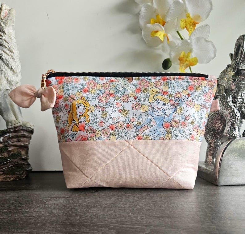 May include: A zippered pouch featuring a top section with a floral pattern and cartoon princess illustrations, and a solid light pink quilted bottom section. A small light pink fabric bow is attached to the side near the black zipper closure.