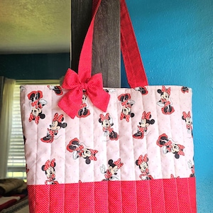 Puede incluir: Un bolso tote acolchado con un estampado de Minnie Mouse sobre un fondo rosa claro. Tiene detalles de lunares rojos, un lazo rojo y correas rojas. El bolso mide aproximadamente 38 cm de ancho y 30 cm de alto.
